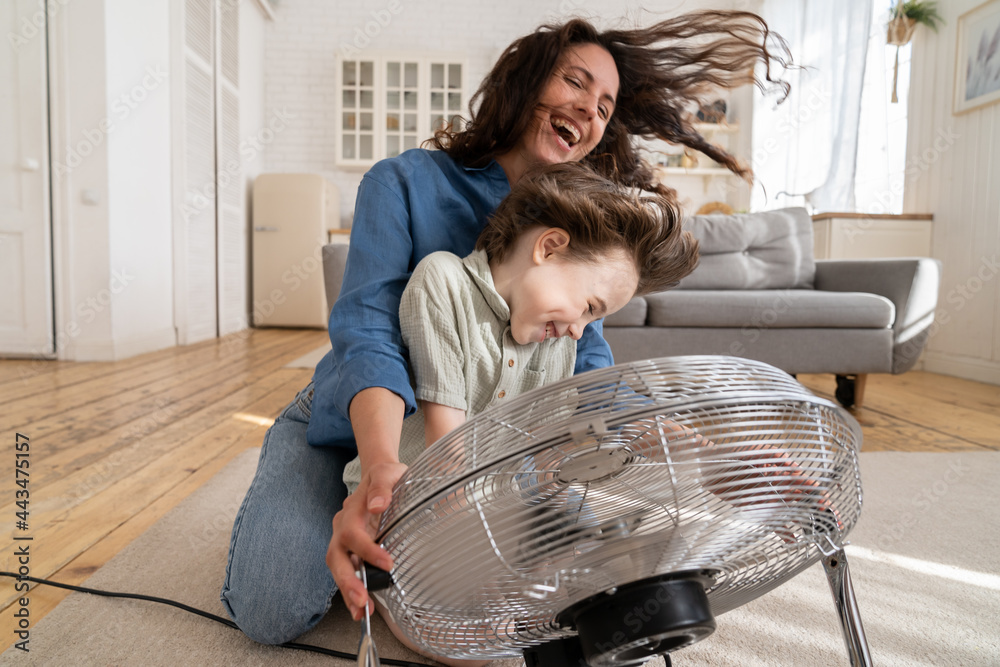 Playful single mother bonding with son excited playing together at home with big fan blowing cool wind in living room. Mum and child enjoy time together at conditioning ventilator during summer heat - obrazy, fototapety, plakaty 