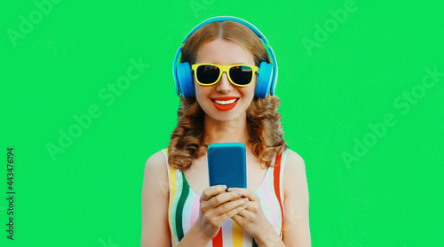 Summer colorful portrait of happy smiling young woman listening to music in headphones with phone on green background