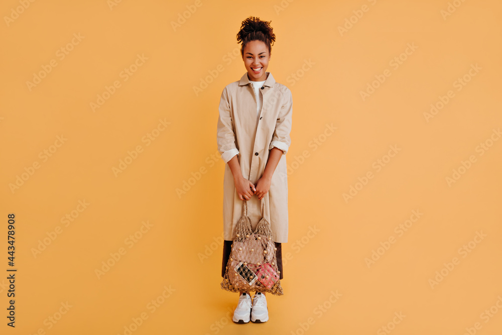 Full length view of fashionable black woman with string bag. Studio shot of african american girl in trench coat looking at camera. Full length view of fashionable black woman with string bag. Studio shot of african american girl in trench coat looking at camera.