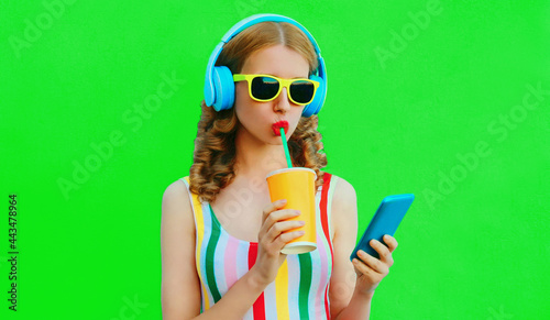 Summer colorful portrait of stylish young woman listening to music in headphones looking at smartphone and drinking fresh juice on green background