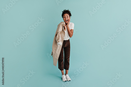 Fashionable black girl holding trench coat. Full length view of excited african american woman laughing on turquoise background.