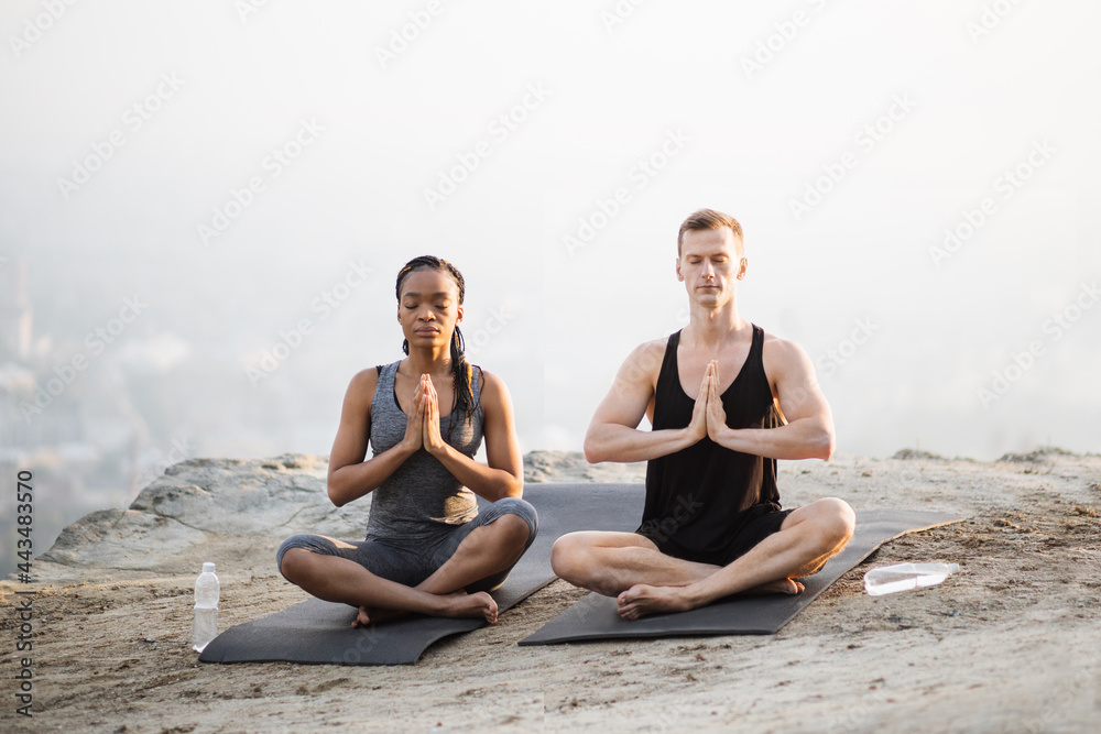 Peaceful multi ethnic couple in sport clothes sitting in lotus position yoga mat and with namaste hands. Young man and woman keeping eyes closed while meditating on fresh air. Peaceful multi ethnic couple in sport clothes sitting in lotus position yoga mat and with namaste hands. Young man and woman keeping eyes closed while meditating on fresh air.