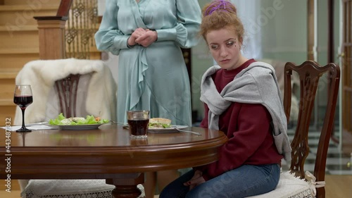 Portrait of bored redhead hipster daughter sitting at table listening to unrecognizable elegant mother scolding. Unhappy Caucasian millennial woman arguing with wealthy parent at home indoors photo