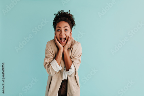 Amazed black lady in trench coat looking at camera with smile. Front view of shocked african american woman posing on turquoise background.