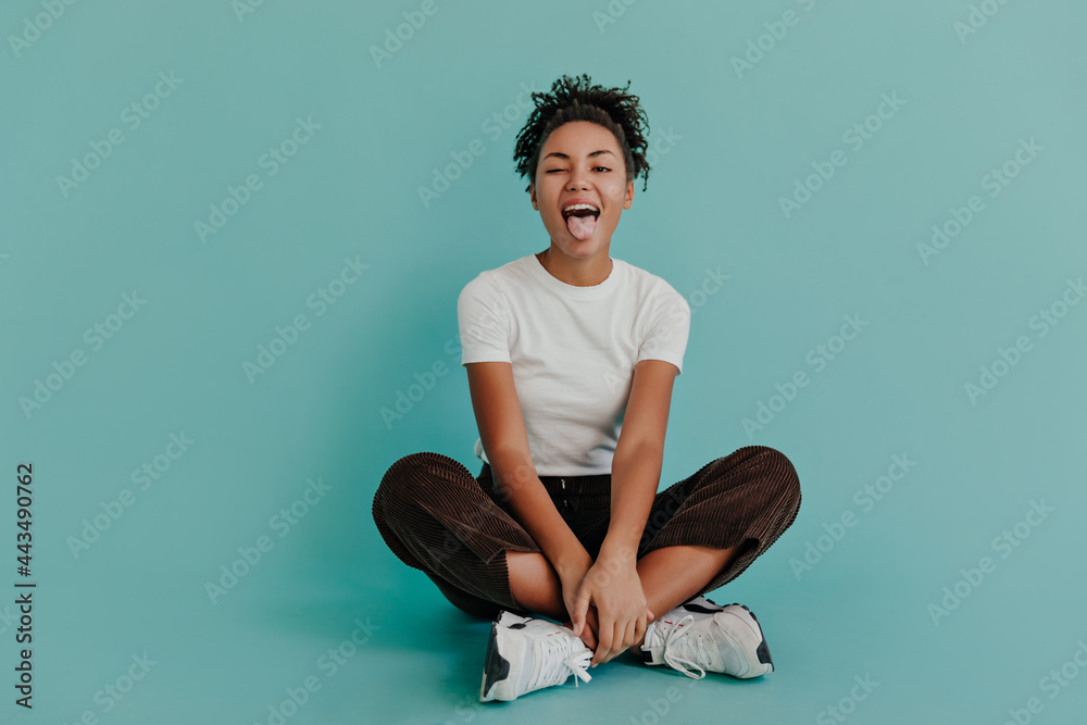 Jocund black girl posing with tongue out on turquoise background. Front view of stunning african american woman sitting with crossed legs.