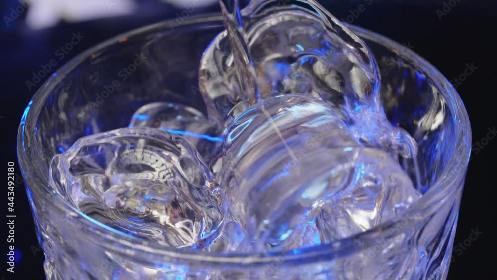 Bartender adds ice cubes to the glass in slow motion, making the cocktail at the bar counter, blue backlight. Bartender adds ice cubes to the glass in slow motion, making the cocktail at the bar counter, blue backlight.