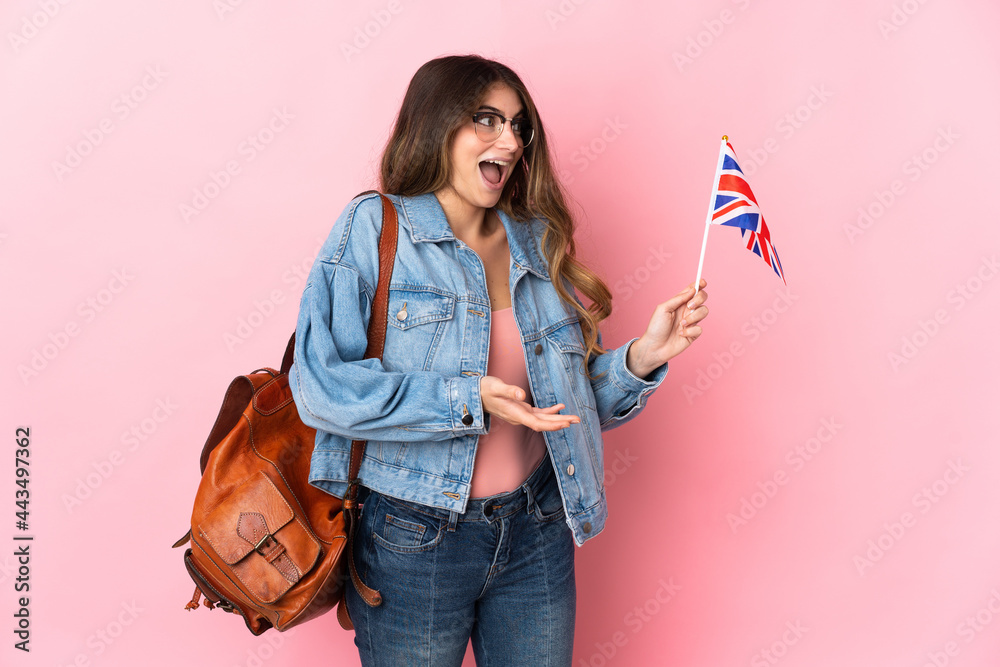 Young woman holding an United Kingdom flag isolated on pink background with surprise expression while looking side