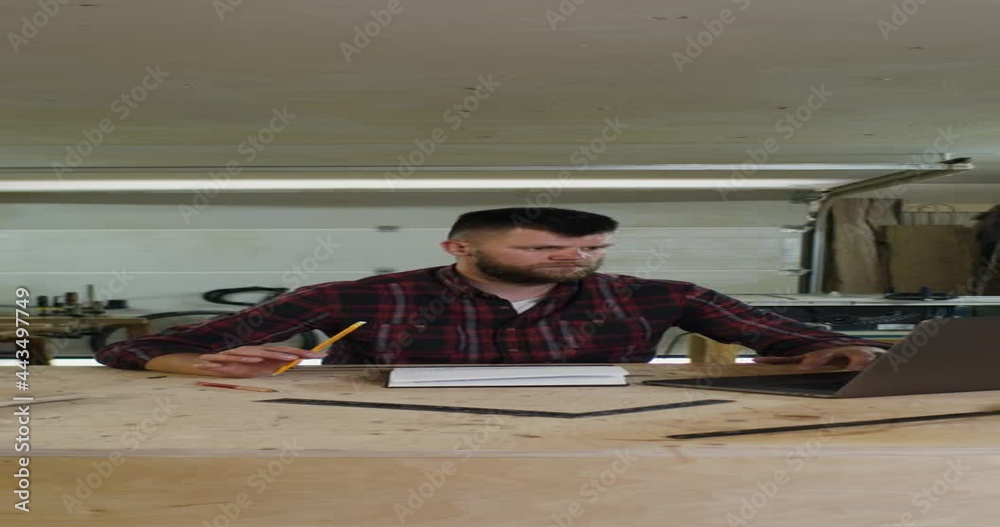 man with beard makes notes in front of open laptop in carpentry workshop man with beard makes notes in front of open laptop in carpentry workshop