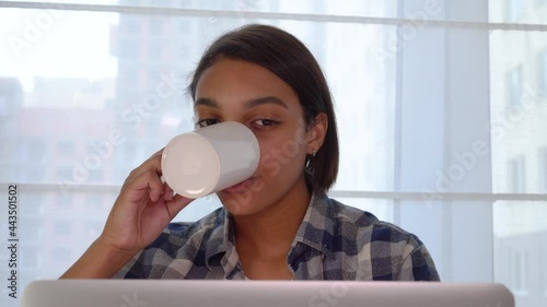 A young woman is looking at a laptop, drinking coffee and chatting with friends online. Safe distance during a pandemic.