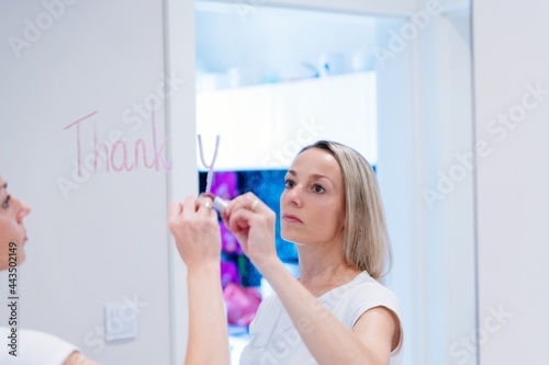 A blonde woman writes the word thank you on the mirror with a lipstick.