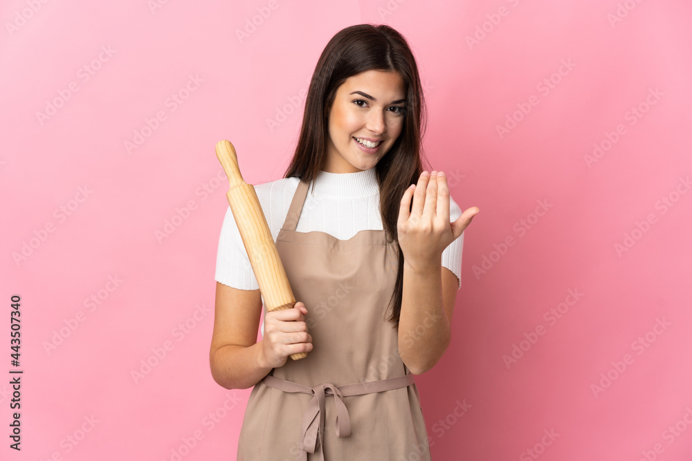 Teenager Brazilian girl holding a rolling pin isolated on pink background inviting to come with hand. Happy that you came