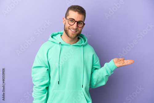 Young handsome caucasian man isolated on purple background holding copyspace imaginary on the palm to insert an ad