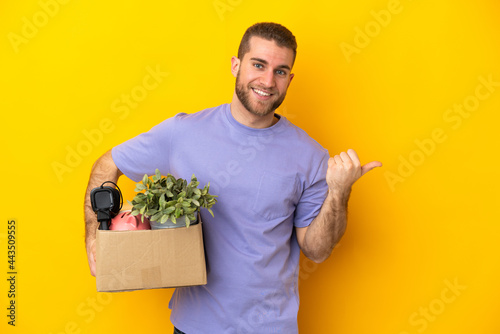 Young caucasian making a move while picking up a box full of things isolated on yellow background pointing to the side to present a product