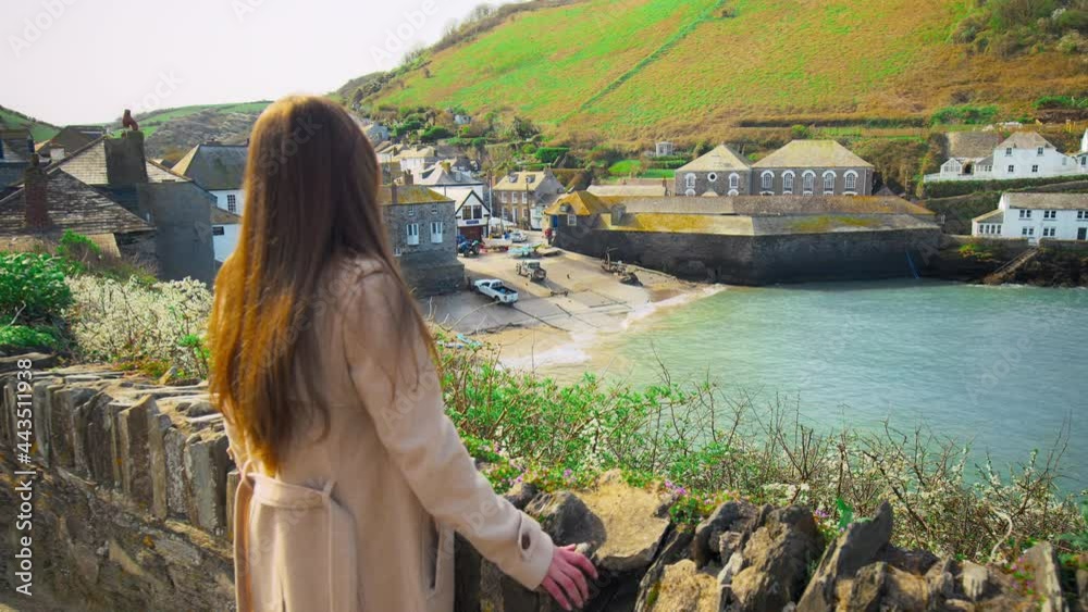 A girl looking at Port Isaac, fishing village, Atlantic coast Cornwall England. A young woman watching at Porthysek, rocky scenic views with blue waters, green lands and old English houses in 4K.