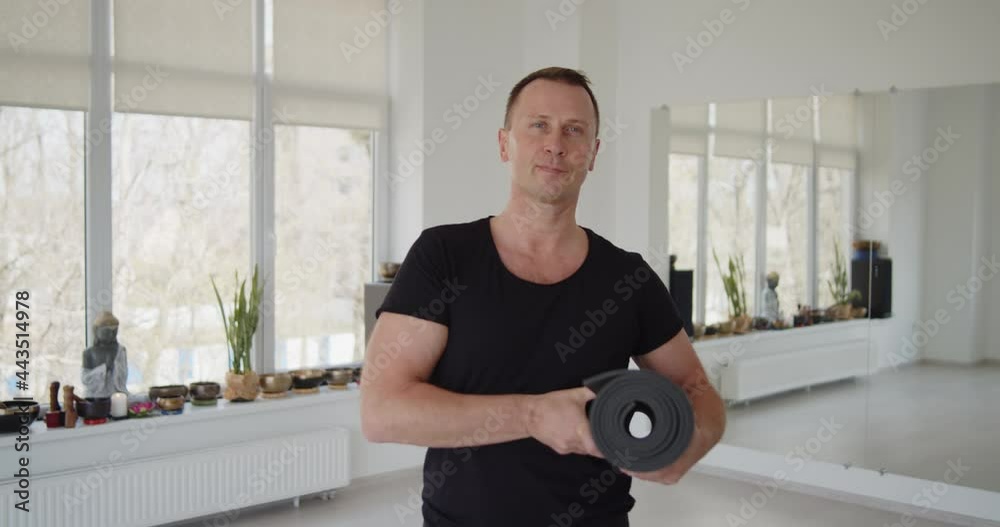 Portrait of a male yoga trainer holding exercise mat standing in a studio looking at the camera. Fitness, healthy lifestyle and wellness concept. Portrait of a male yoga trainer holding exercise mat standing in a studio looking at the camera. Fitness, healthy lifestyle and wellness concept.
