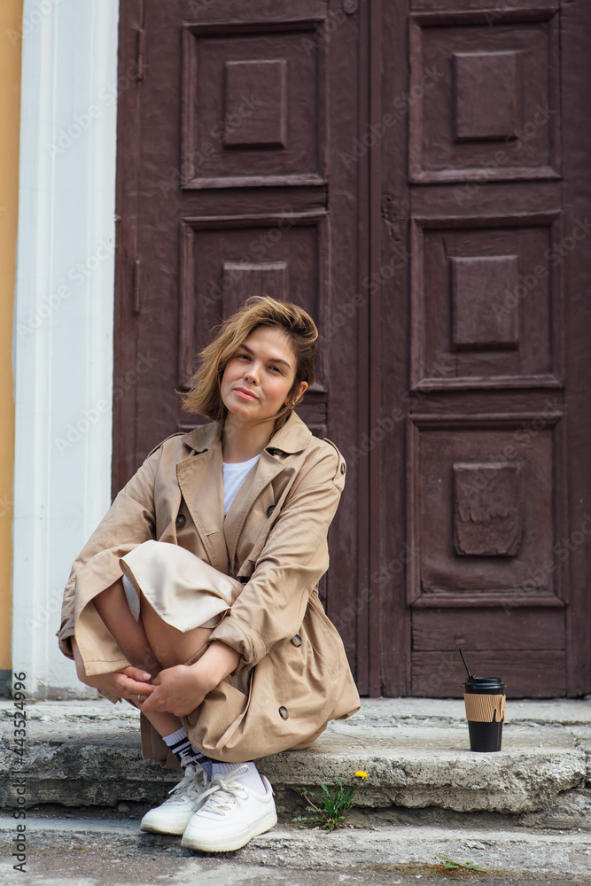 Young millennial woman with wild hair dressed in an autumn coat sitting near the door of an old building with coffee to go.