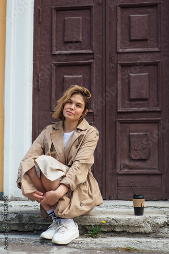 Young millennial woman with wild hair dressed in an autumn coat sitting near the door of an old building with coffee to go.