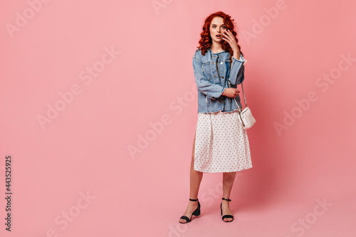 Full length view of shocked ginger woman in high-heeled shoes. Studio shot of amazed girl in denim jacket standing on pink background.