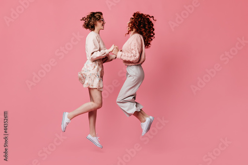 Two female friends holding hands and looking at each other. Side view of amazing girls jumping on pink background.