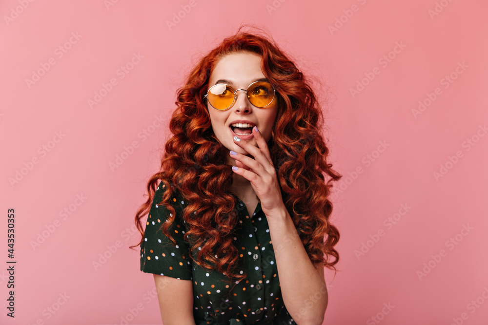 Dreamy ginger girl looking away with open mouth. Studio shot of emotional young woman in sunglasses posing on pink background.