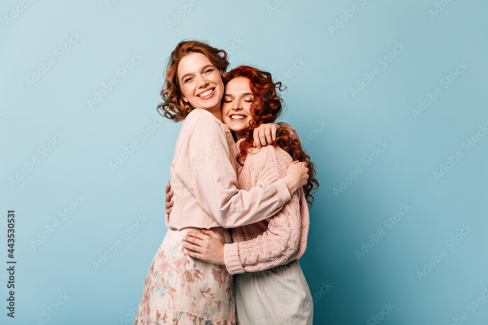 Debonair girls embracing on blue background. Studio shot of attractive friends laughing at camera. Debonair girls embracing on blue background. Studio shot of attractive friends laughing at camera.