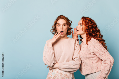 Shocked ladies sharing rumors. Studio shot of amazed gossip girls posing on blue background.