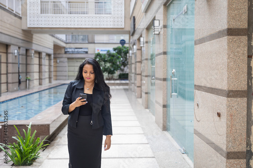 An Indian woman standing looking into her phone in an urban corporate setting. Corporate female employee in smart formal attire