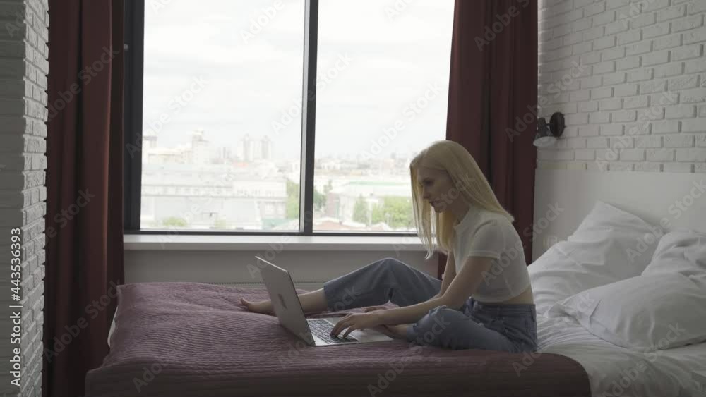 Young woman works at a laptop in a cozy bright bedroom. The blonde woman is typing on the keyboard against the background of a large window. Remote work concept. 4K UHD Young woman works at a laptop in a cozy bright bedroom. The blonde woman is typing on the keyboard against the background of a large window. Remote work concept. 4K UHD