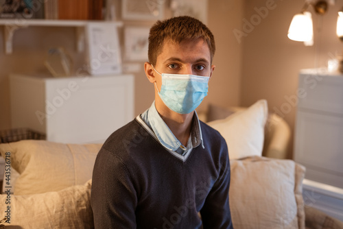 a young guy of Caucasian ethnicity in a shirt and a protective mask sits on the couch alone at home. Concept of being at home during global quarantine
