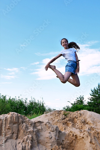 cute cheerful girl makes a jump in air against background of blue sky. Female athlete in white T-shirt and denim shorts performs acrobatic exercises. Copy space