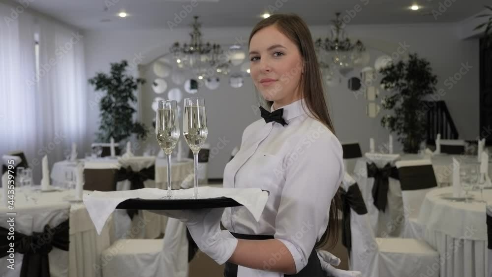 A young smiling female waiter holds a tray of champagne in a restaurant.