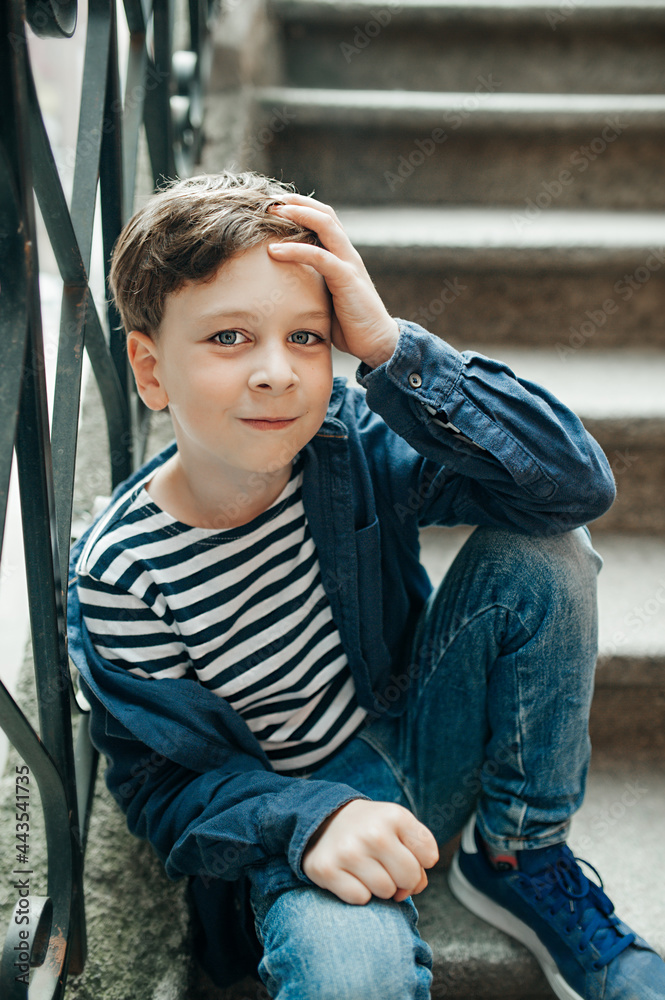 stylish fashionable schoolboy boy in vest and blue shirt is sitting on stairs in fashionable pose on city street. portrait of beautiful boy. vertical, selective focus