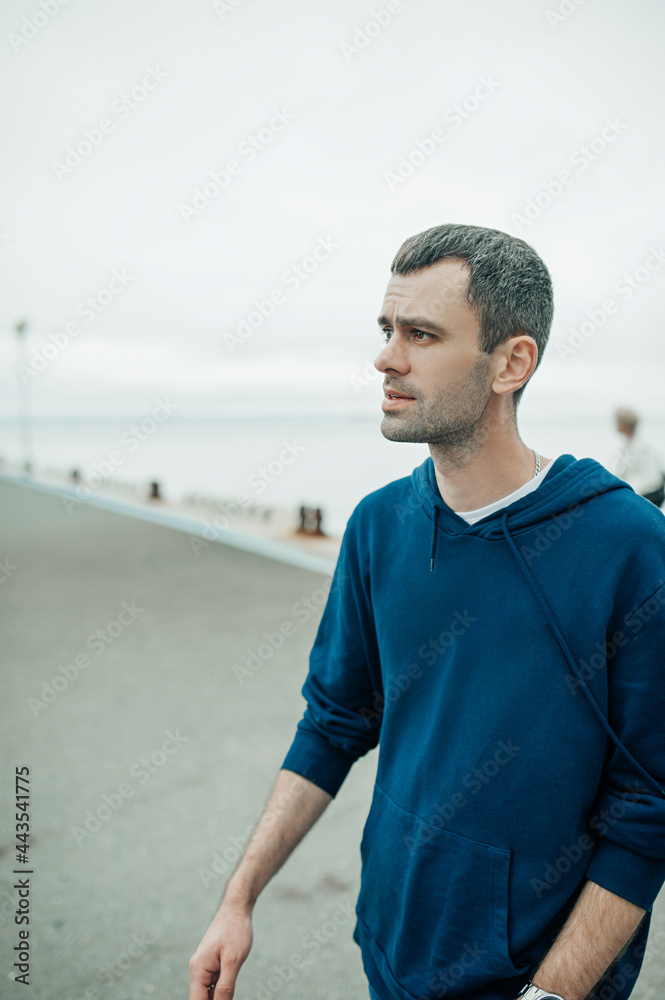 Obraz premium middle-aged man walks on the sea pier. portrait of young man with dark but already partially gray hair in blue hoodie near the sea. urban style content. selective focus