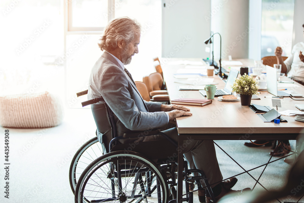 Portrait of sad disabled businessman sitting in wheelchair in office with colleagues working on background. Handicapped lonely male employee in wheelchair sitting in office Portrait of sad disabled businessman sitting in wheelchair in office with colleagues working on background. Handicapped lonely male employee in wheelchair sitting in office