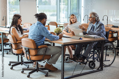 Large group of colleagues during a presentation in board room. Focus is on man in wheelchair. One Businessman on a wheelchair sitting at the table and discussing with colleagues in office