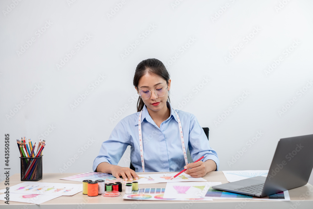 Beautiful young Asian fashion tailor with textile sewing accessories and entrepreneur designer sketches are full of bright colors on the desk with a laptop for creative ideas in the studio Beautiful young Asian fashion tailor with textile sewing accessories and entrepreneur designer sketches are full of bright colors on the desk with a laptop for creative ideas in the studio