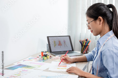 Beautiful young Asian fashion tailor with textile sewing accessories and entrepreneur designer sketches are full of bright colors on the desk with a laptop for creative ideas in the studio