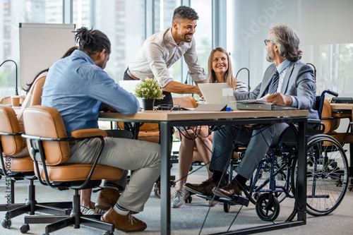 A man in a wheelchair is working in a bright office. His colleagues are passionate about work. Young businessman greeting handicapped business partner and team. Coworker on wheelchair