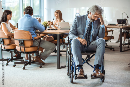 Disabled businessman in wheelchair covering face with hands in office. Man in a wheelchair holding his head in hands and feeling frustrated while sitting at his workplace in the office