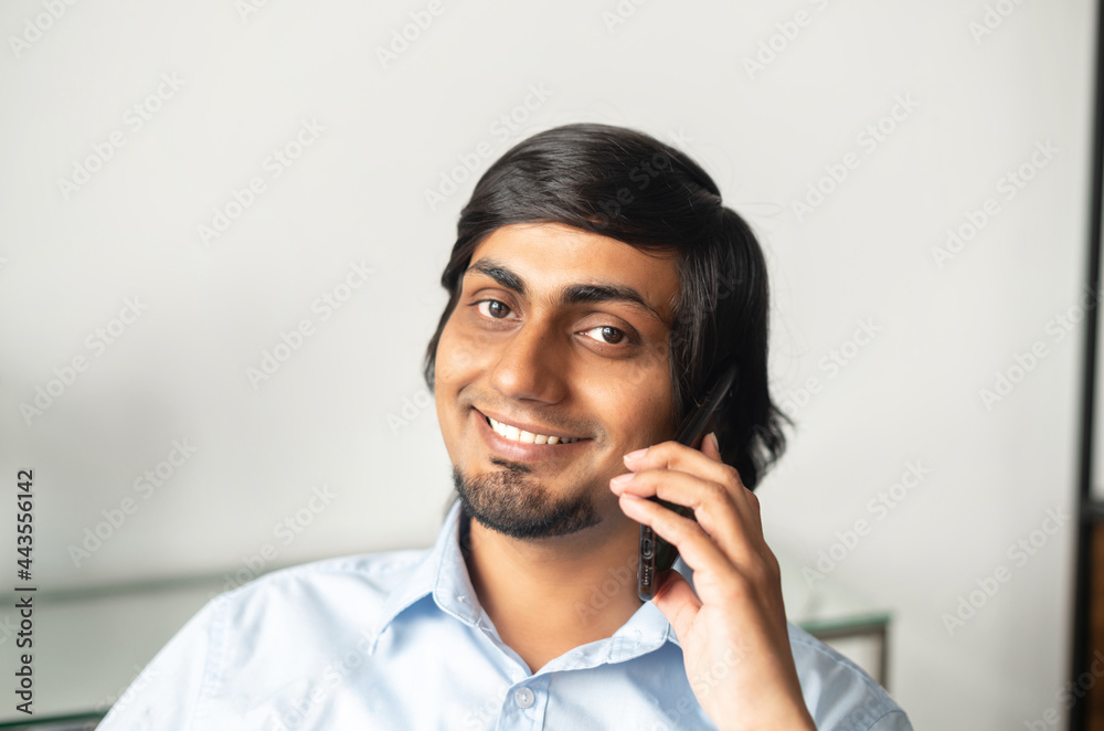 Headshot of smiling indian businessman in smart casual shirt talking on the smartphone and looks at the camera, eastern man has pleasant phone conversation Headshot of smiling indian businessman in smart casual shirt talking on the smartphone and looks at the camera, eastern man has pleasant phone conversation