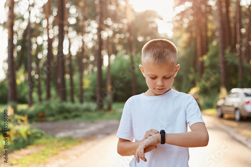 Little blonde boy using fitness bend touching button and touchscreen while posing outdoor in park, touching button and setting smart watch before running on road. photo