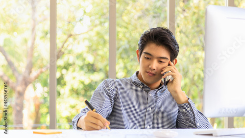 Business and Comminication Concept. Asian young businessman make a phone call by smartphone and take a note on paper in office with desktop computer. photo