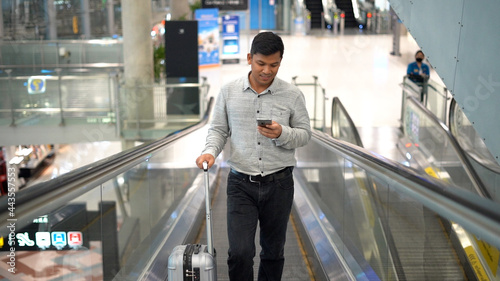 Young man with suitcase standing on the moving walkway while using smartphone.