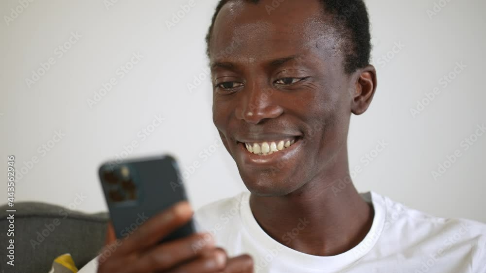 Thrilled young afro-american guy in a white t-shirt looking at the mobile phone and smiling for a message, good news or a win. 