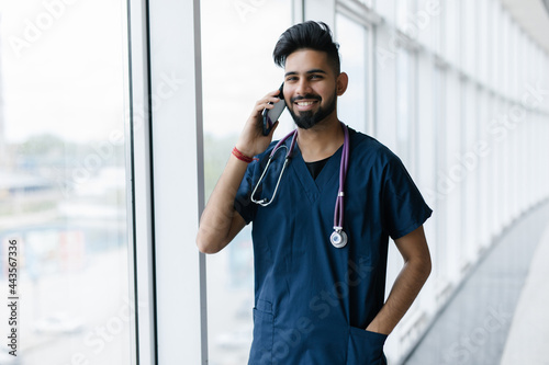 Young Indian Doctor in White Uniform with Phone. Consulting Doctor. Medical Staff in Clinic. Bearded Man with Smartphone. Healthcare Professional Concept. Young Man with Stethoscope.