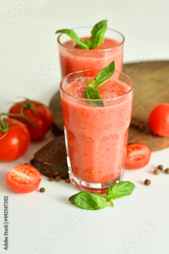 Two glasses of fresh organic tomato juice decorated with raw tomatoes and green leaves of basil on light background.Healthy diet nutrition concept.