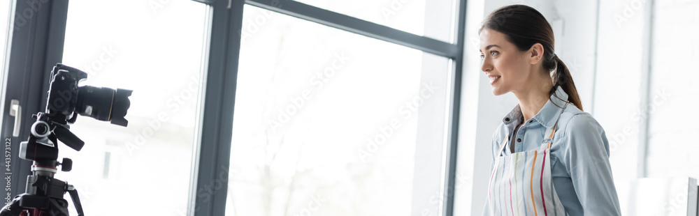 smiling housewife in apron talking near digital camera and window at home, banner. smiling housewife in apron talking near digital camera and window at home, banner.