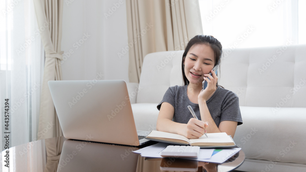 Asian female talking on the phone and working in a laptop with a smiling face, Business Cell Phone Conversations, Write notes in a notebook as a reminder, Working at home, Home lifestyle, Stay home. Asian female talking on the phone and working in a laptop with a smiling face, Business Cell Phone Conversations, Write notes in a notebook as a reminder, Working at home, Home lifestyle, Stay home.
