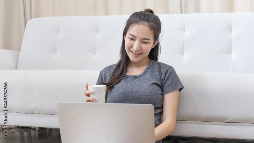 Asian woman sitting on laptop and sipping coffee in living room on weekends, Entertaining or relaxing using laptop technology, Wake-up activities, New work lifestyle, Happy time, Living at home. Asian woman sitting on laptop and sipping coffee in living room on weekends, Entertaining or relaxing using laptop technology, Wake-up activities, New work lifestyle, Happy time, Living at home.