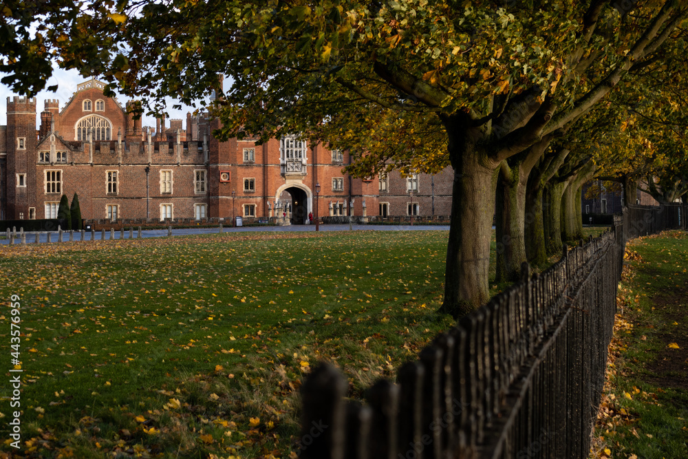 Bright red Tudor Facade of Hampton Court Palace seen through trees that are alongside the river Thames footpath side